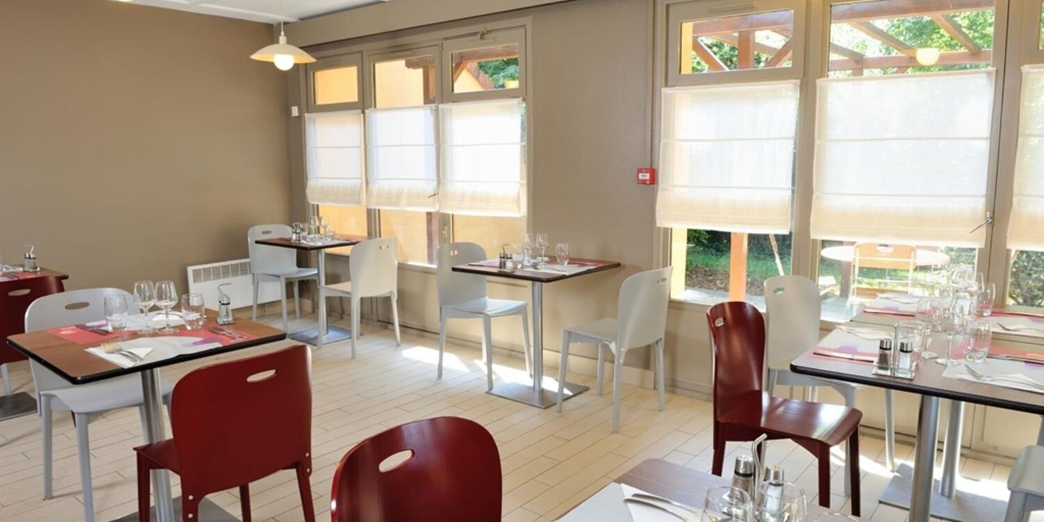 Salle du restaurant avec tables dressées, chaises blanches et rouges, baies vitrées sur le jardin